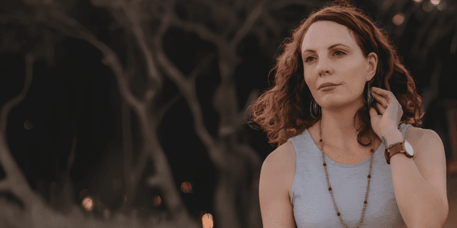A woman with curly hair wearing a sleeveless top, gazing thoughtfully outdoors
